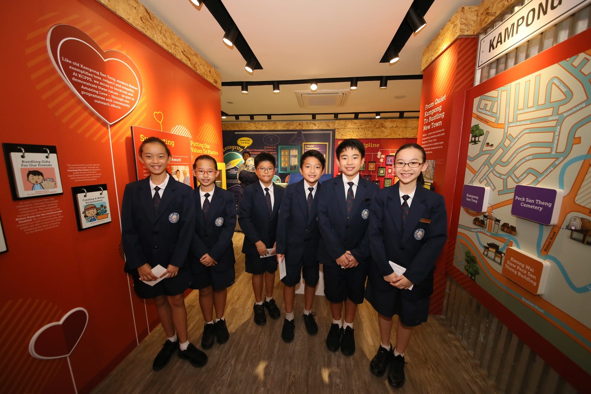 Six students in navy blue uniforms smile inside a colorful school exhibition hall with orange walls and maps.